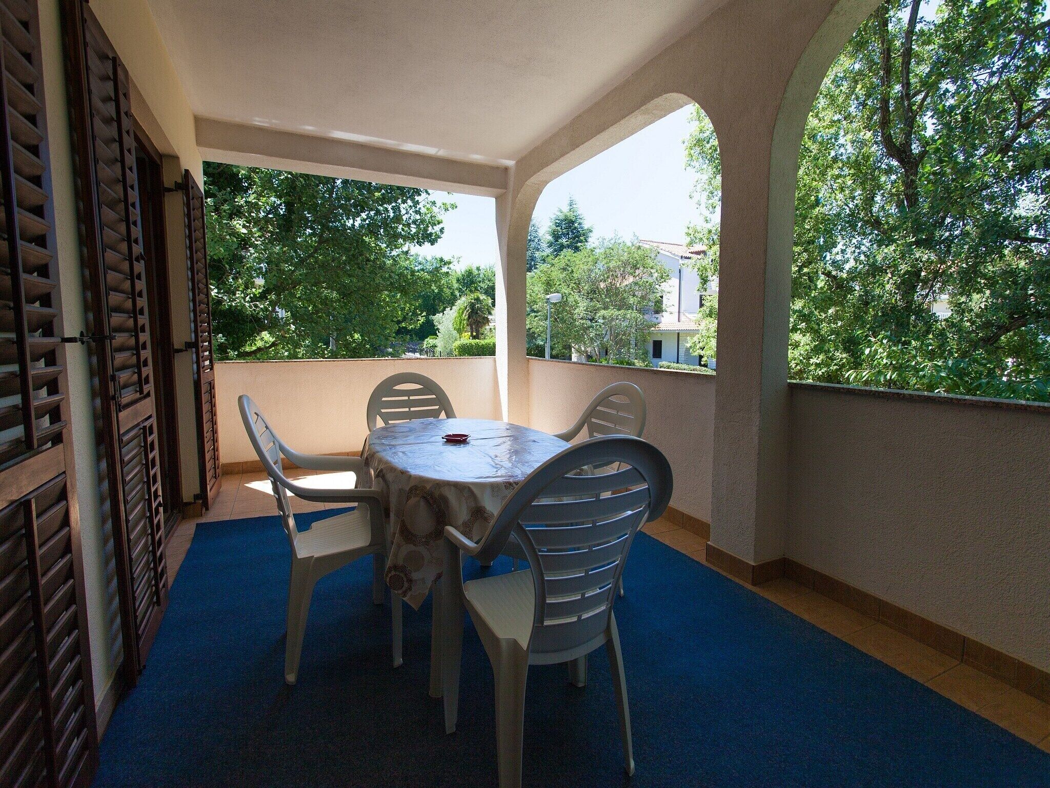 Un espacio de estar al aire libre techado con una mesa y sillas, alfombra azul y vistas a árboles y edificios.