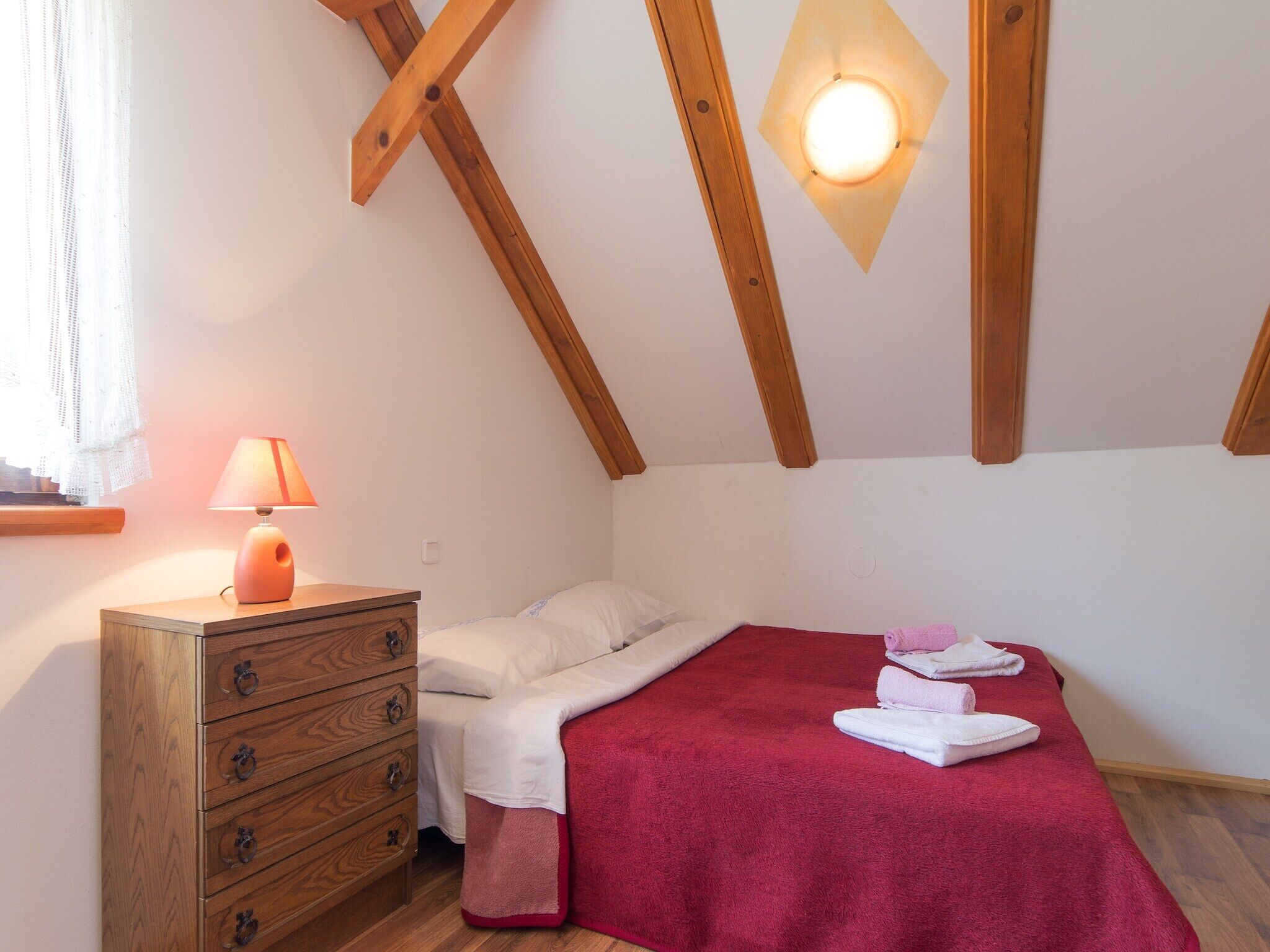 Une chambre à coucher avec une commode en bois, un lit recouvert d’une couverture rouge et une table de chevet avec une lampe.