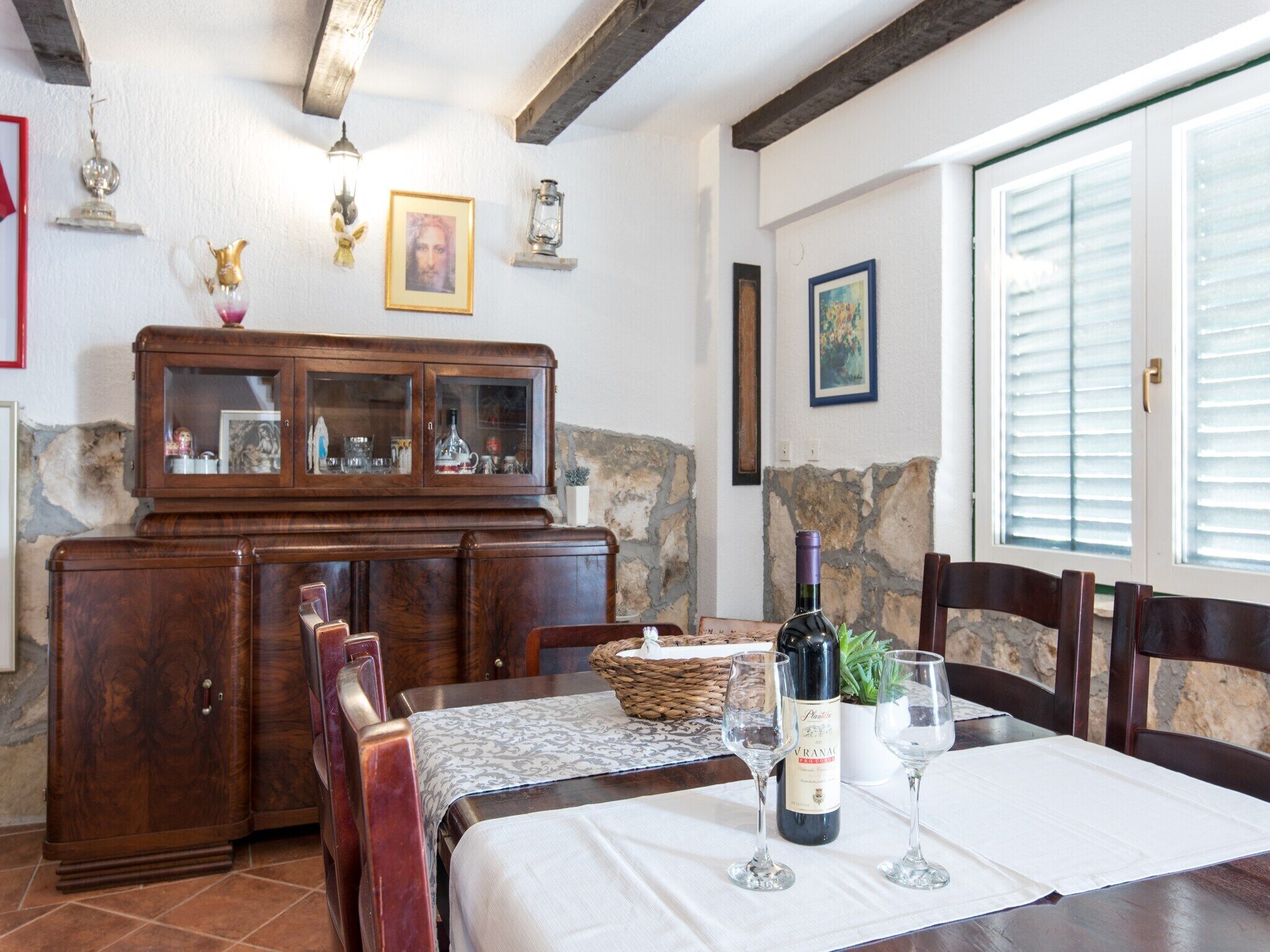 A dining area with a wooden table set for two, a wine bottle, and two wine glasses. There is a cabinet with glass doors and a stone wall in the background.