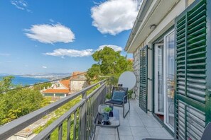 A balcony with a view of the sea, a table with chairs, and a window with shutters.