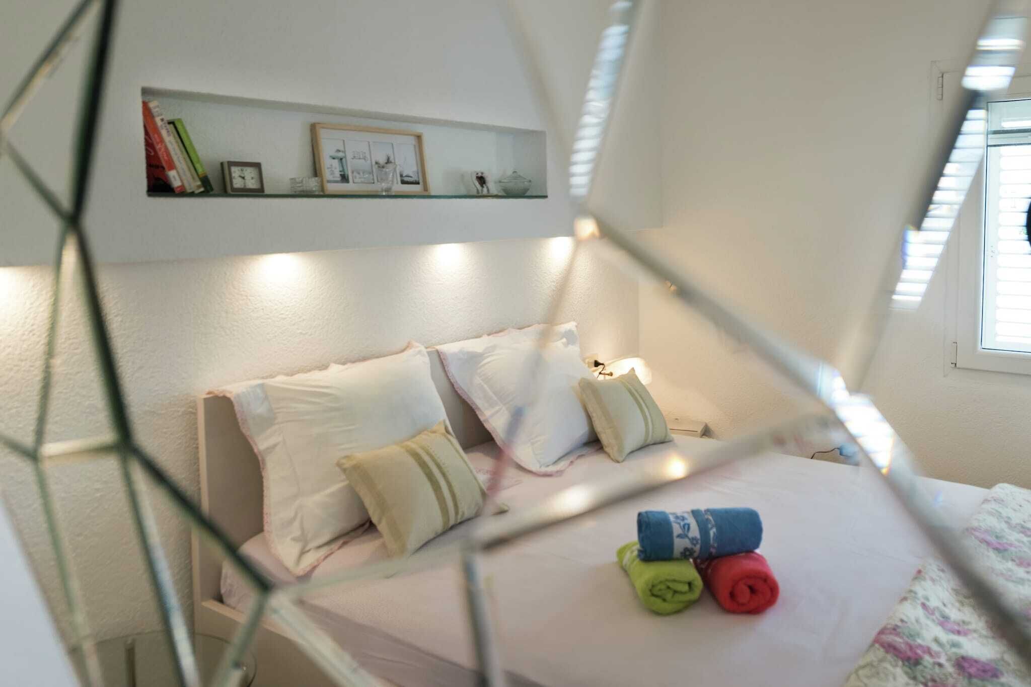 A cozy bedroom with a bed, pillows, a small shelf with books and a clock, and a window with blinds.