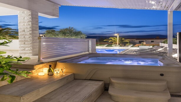 Uma banheira de hidromassagem com degraus e área de estar, rodeada por um alpendre coberto com vista para uma piscina e edifícios ao fundo.