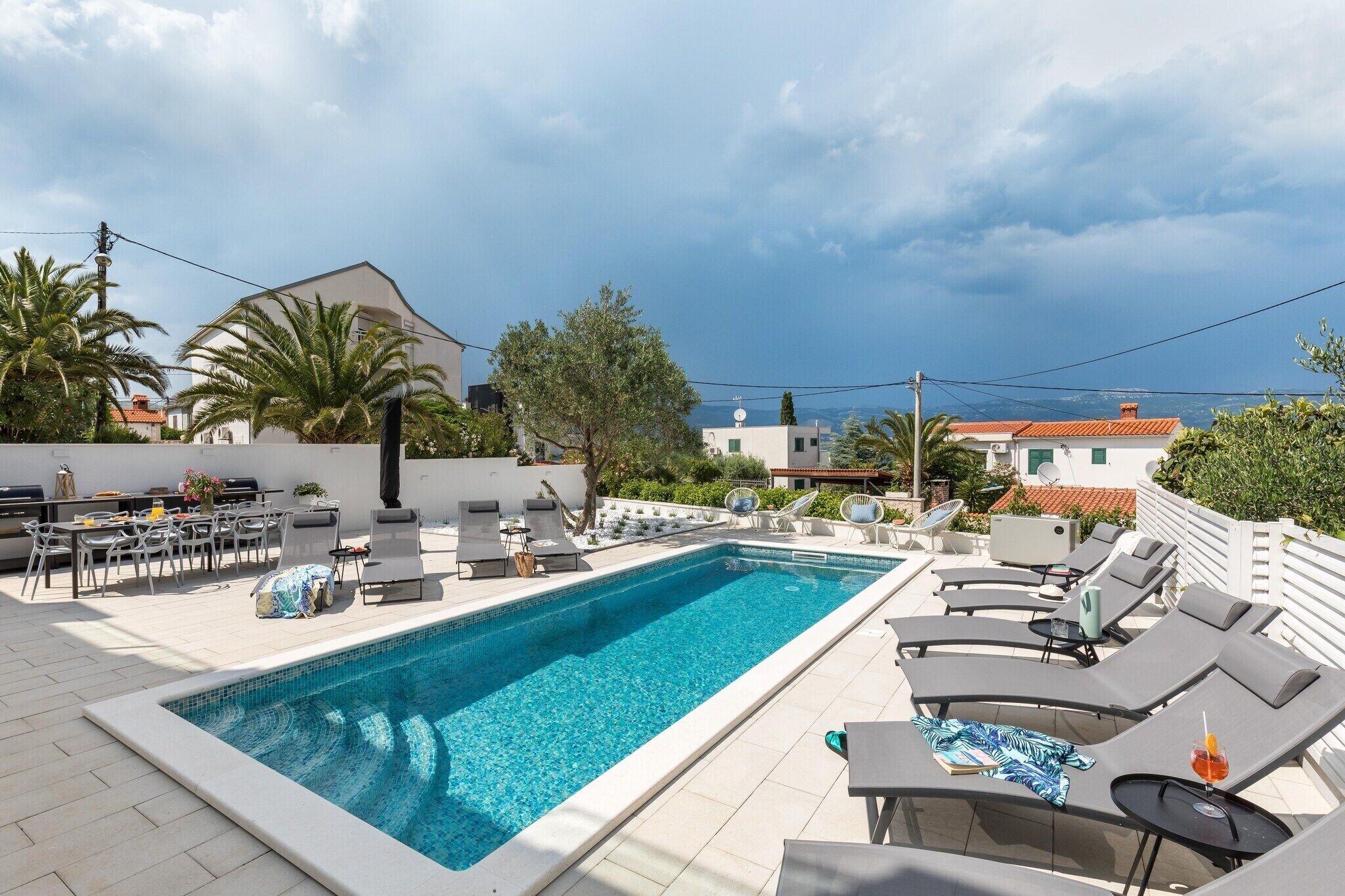 A rooftop pool area with lounge chairs and a dining table, surrounded by greenery and overlooking a coastal landscape.