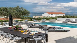 Un espace aménagé au bord de la piscine, avec des chaises longues, une table à manger où sont servis les articles du petit-déjeuner, et une vue sur la mer et un bâtiment en arrière-plan.