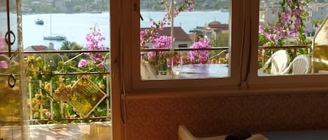 Una habitación con cama, ventana con vistas al agua y barcos, y balcón con flores.