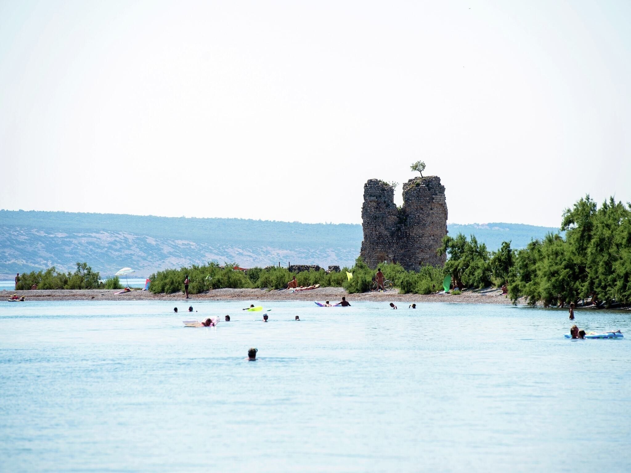 A coastal area with a prominent rock formation in the background, a beach with people swimming and sunbathing, and a clear view of the distant shoreline.