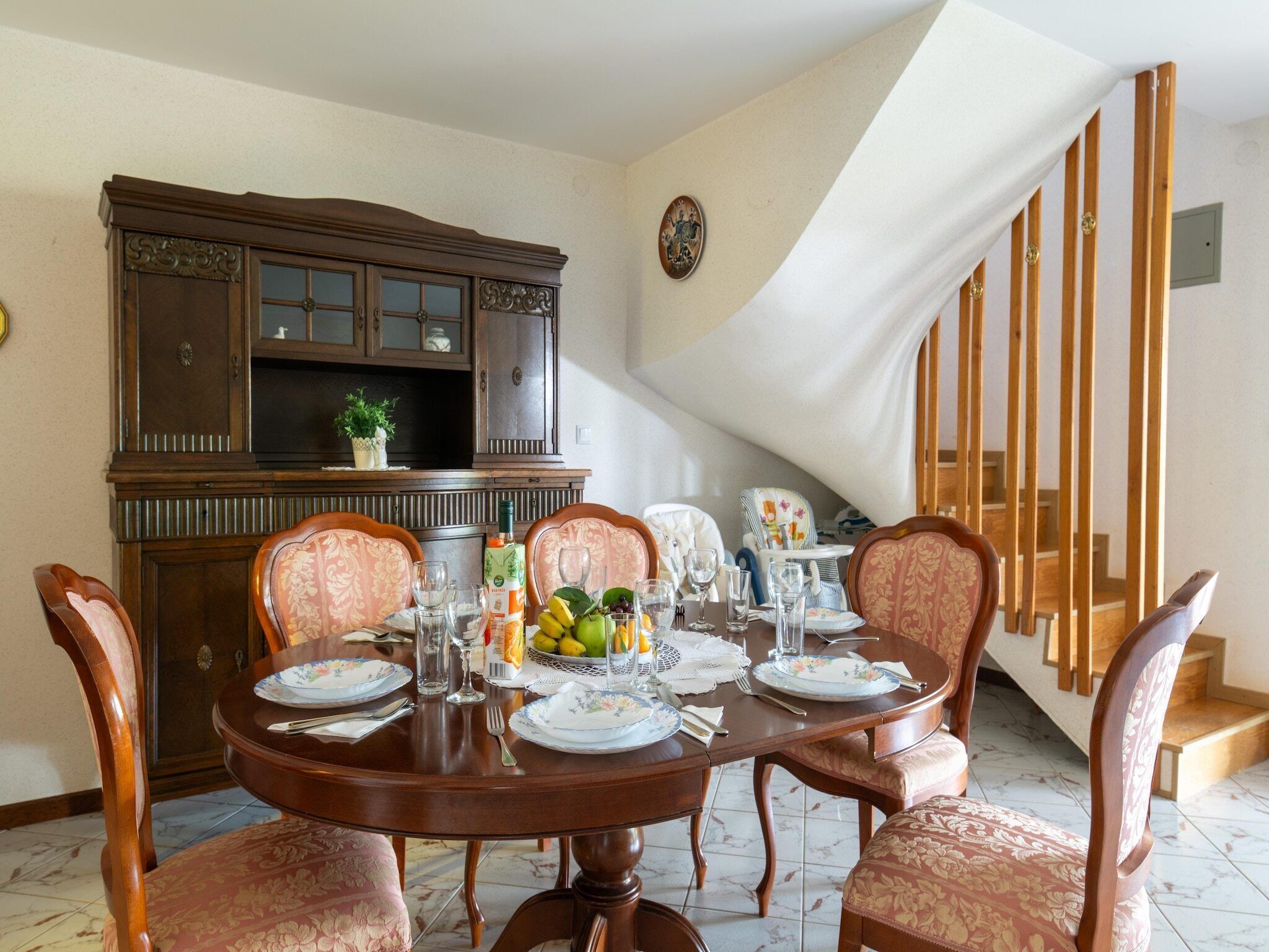 Un coin repas avec une table ronde dressée pour quatre personnes, entourée de chaises à motifs floraux. Un buffet en bois avec des portes en verre se trouve à l’arrière-plan, et un escalier avec des rampes en bois est visible sur la droite.