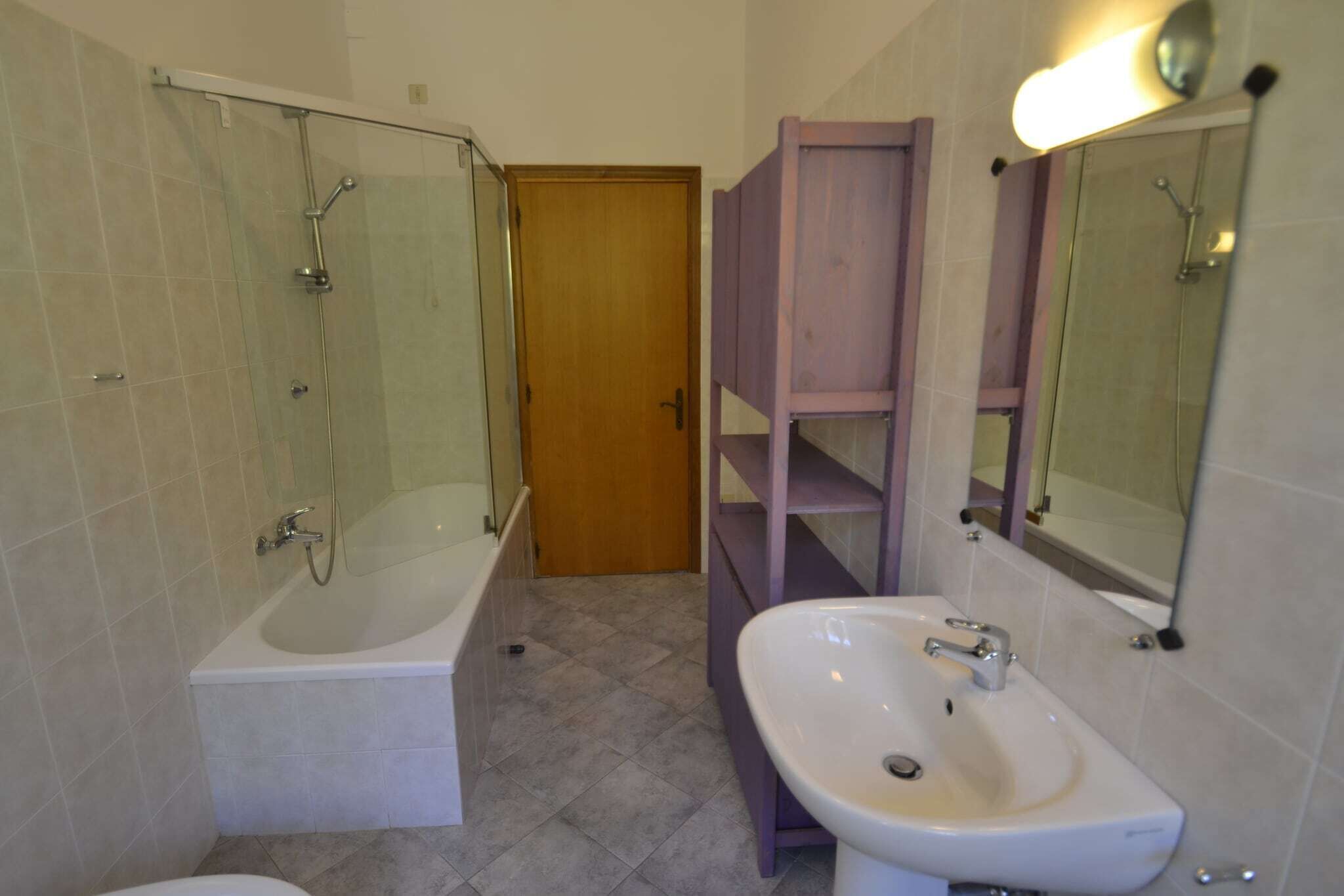 A bathroom with a glass-enclosed shower, a white bathtub, a sink with a mirror, and a wooden shelf.