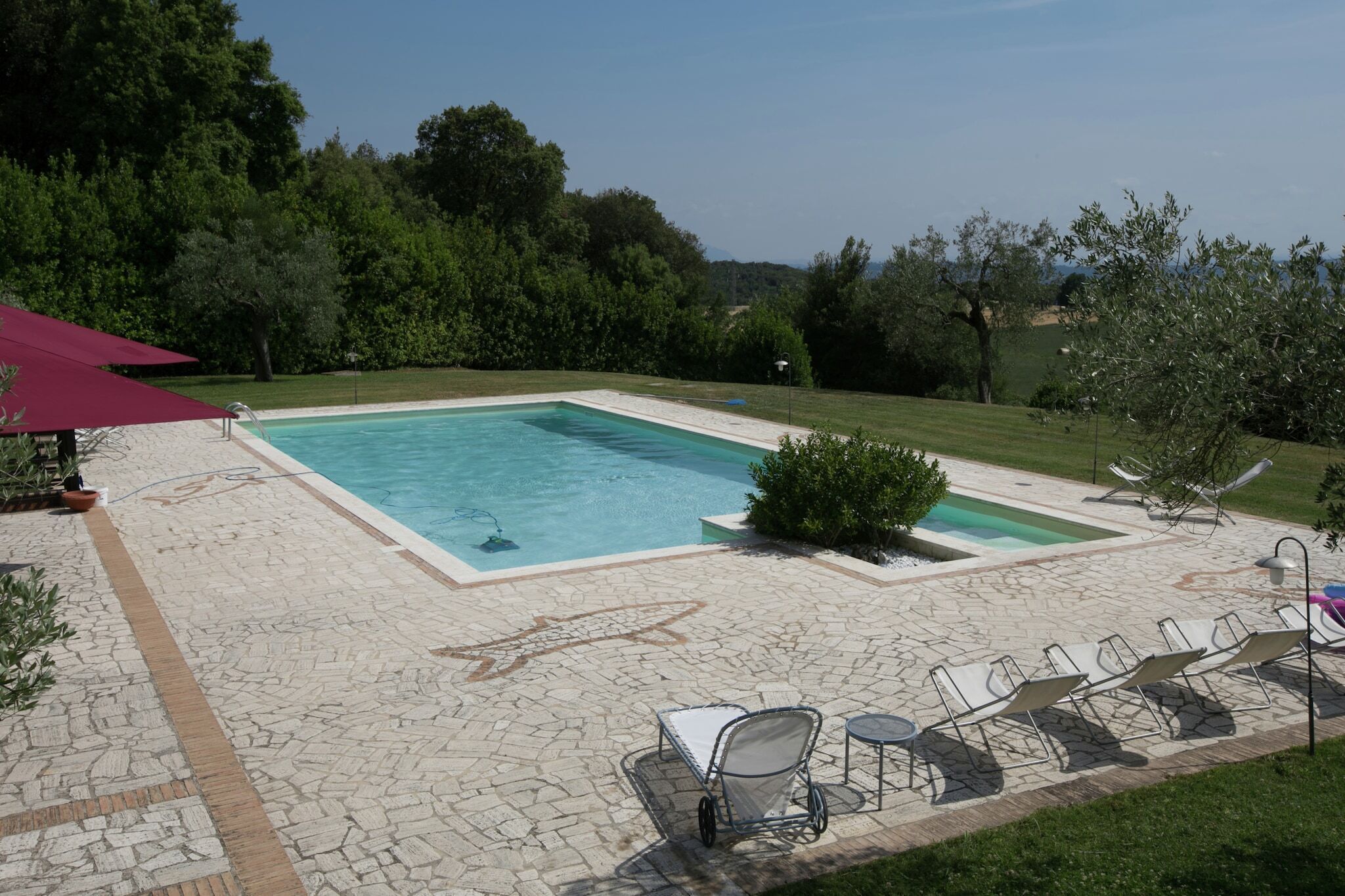Une piscine rectangulaire, bordée d’une terrasse carrelée, entourée d’une pelouse et d’arbres.