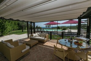 Un espacio de descanso al aire libre con mobiliario de mimbre, una mesa de vidrio con bebidas y vistas a una piscina y montañas.