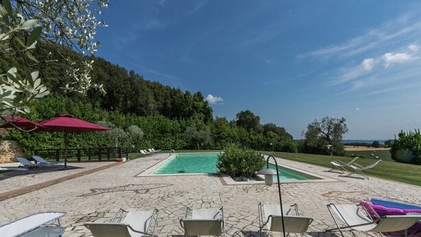 Ein Poolbereich mit Liegestühlen und Sonnenschirmen, umgeben von einer Steinterrasse und Grünflächen.