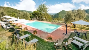 Piscina rectangular con una solana circundante, rodeada de sillas de descanso y sombrillas, con colinas y árboles al fondo.