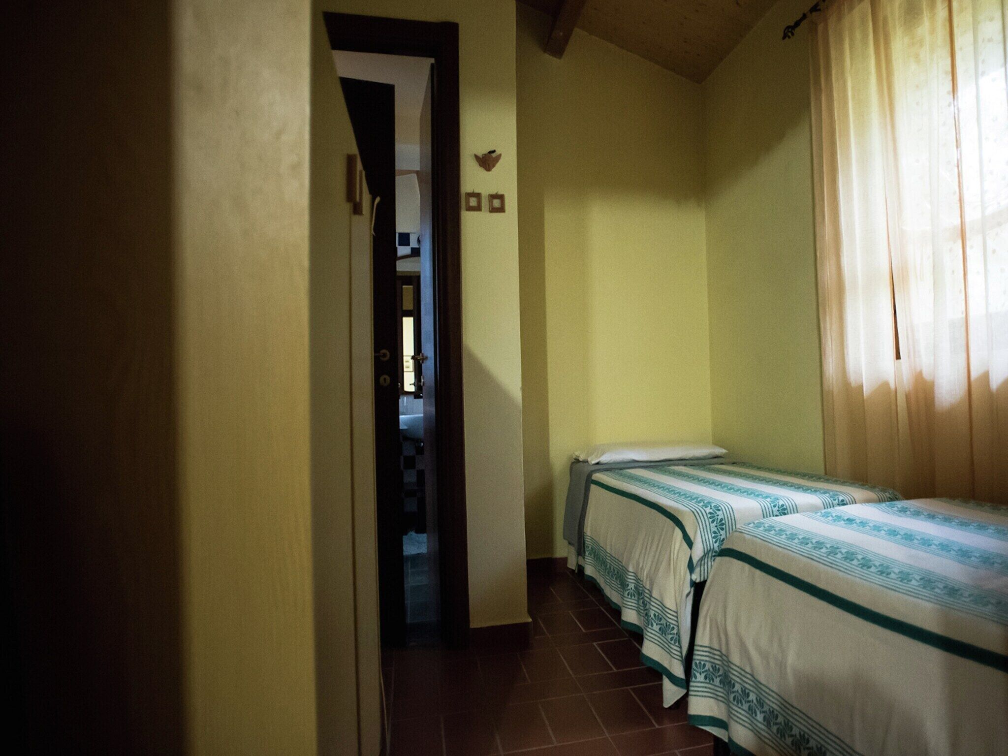 A narrow hallway leading to a room with two beds, a door, and a small window with curtains.