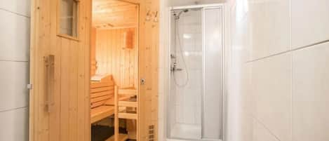 Una sauna de madera con un banco y un balde pequeño, junto a un área de ducha con paredes de vidrio.