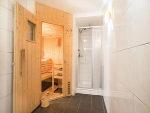 Eine Holzsauna mit einer Bank und einem kleinen Eimer, neben einem vom Glas umschlossenen Duschbereich.
