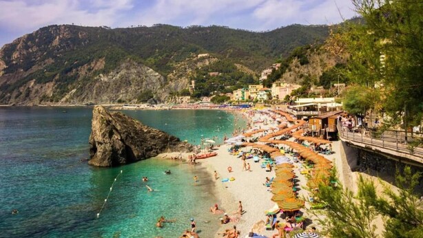 Une plage côtière aux eaux cristallines, avec un promontoire rocheux et des bâtiments qui bordent le rivage.