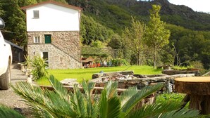 Un bâtiment en pierre avec une partie supérieure blanche et une porte verte, entouré d’une végétation luxuriante et adossé à un paysage montagneux.