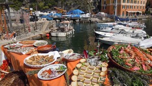 Une table dressée avec des fruits de mer et du pain, située au bord de l’eau, avec des bateaux et des bâtiments en arrière-plan.