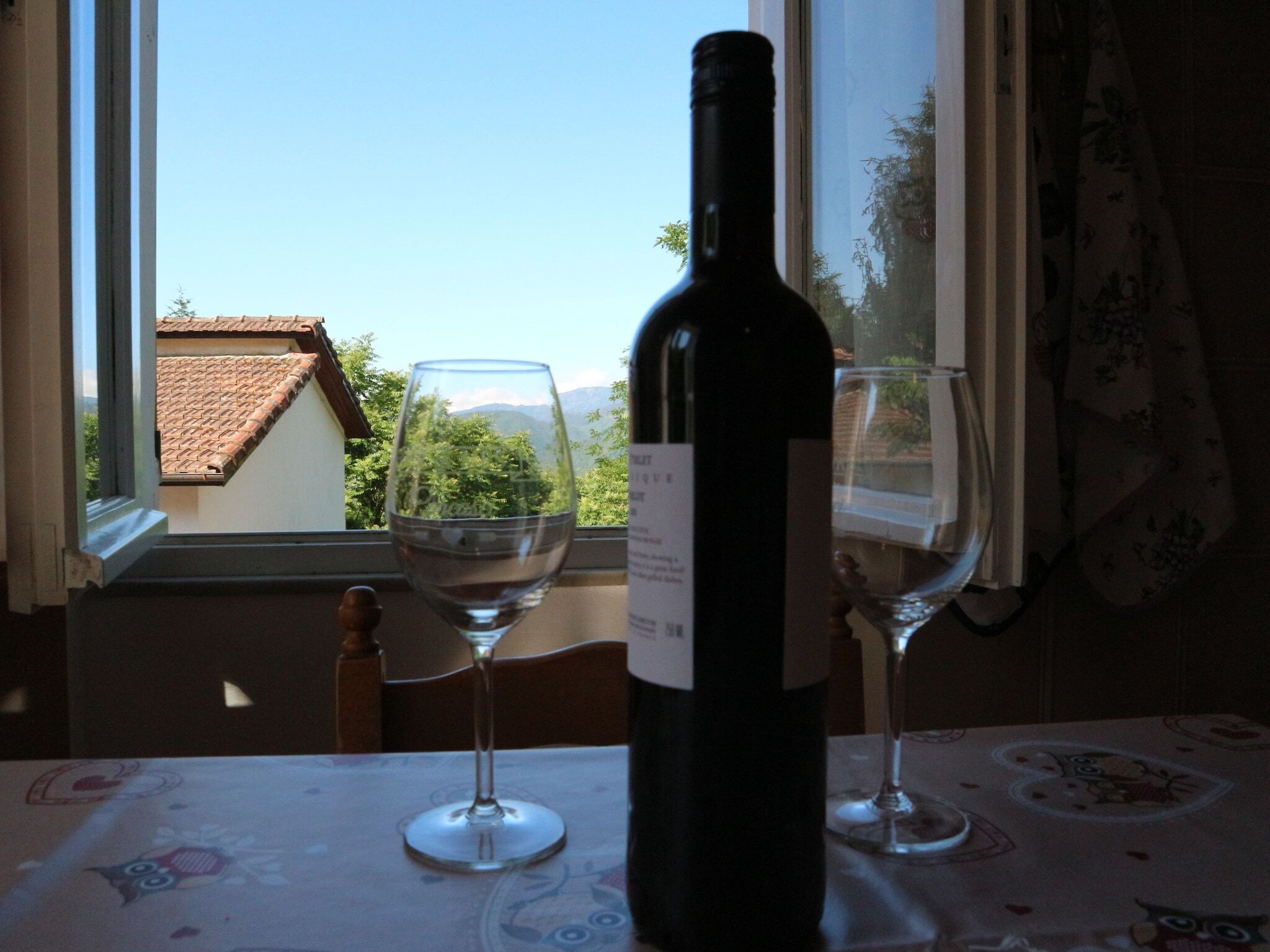 Una botella de vino y dos copas sobre una mesa, cerca de una ventana con vistas a edificios y árboles.