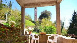 A covered outdoor seating area with two white plastic chairs, surrounded by potted plants and a view of a clock tower in the background.