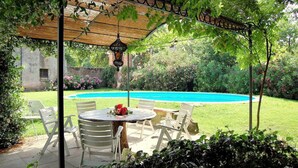 Un patio cubierto con piscina, sillas blancas y una mesa con un centro de mesa de flores rojas.