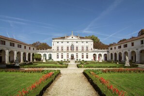 Un edificio grande y simétrico con una entrada central, flanqueada por dos alas, rodeado de un jardín formal con un camino de grava que lleva a la entrada.