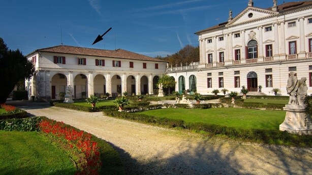 Un edificio grande con múltiples arcos y un patio con estatuas y flores rojas.