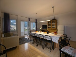 Een moderne woonkamer met een bank, een eettafel met stoelen, een keuken met een kast en een koelkast, en een groot raam met uitzicht op de omgeving.