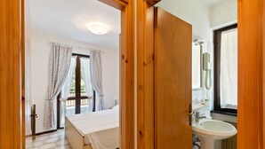 A bedroom with a bed, a window with curtains, a bathroom with a sink and mirror, and a wooden door. - Apartment in Ledro by Lake & Dolomites (Ledro)