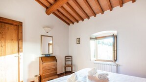 A bedroom with a wooden dresser, a chair, a bed with towels, a window, and a radiator.