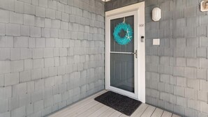 A corner of a building with grey brick walls, a white door with a glass panel, and a teal wreath hanging on the door.