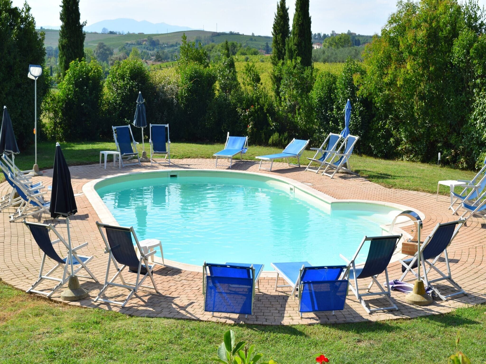 A swimming pool with blue chairs and umbrellas, surrounded by greenery and hills in the background.