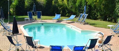 A swimming pool with blue chairs and umbrellas, surrounded by greenery and hills in the background.
