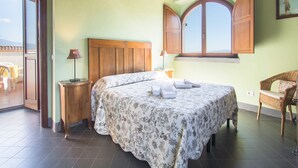 Ein Schlafzimmer mit Bett, Holz-Kopfstück, Fenster mit Holzläden, einem Korbstuhl und Blick ins Freie.