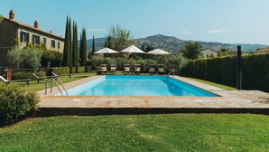 Una piscina rectangular con agua cristalina azul, rodeada por un césped cuidado y un sendero de piedra. Hay sillas de descanso y sombrillas para relajarse, y un edificio de piedra con contraventanas verdes al fondo.