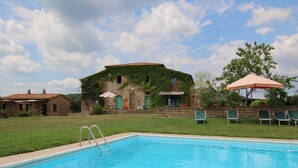 Una piscina con un edificio de piedra al fondo, una zona ajardinada y algunas sillas de descanso.