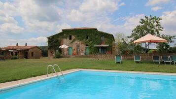 Una piscina con un edificio in pietra sullo sfondo, una zona verde e alcuni lettini.