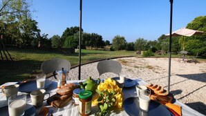 Un área de comedor al aire libre con una mesa preparada para dos personas, mostrando una variedad de comida y bebidas, una tetera verde y un arreglo floral amarillo.