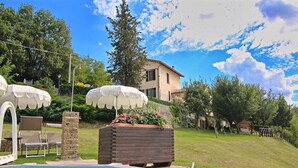 A swimming pool with a tiled edge, surrounded by a grassy lawn and a wooden deck. There are two white umbrellas and a planter with flowers. - Flat in Piticchio with Scenic Views & Pool (Arcevia)