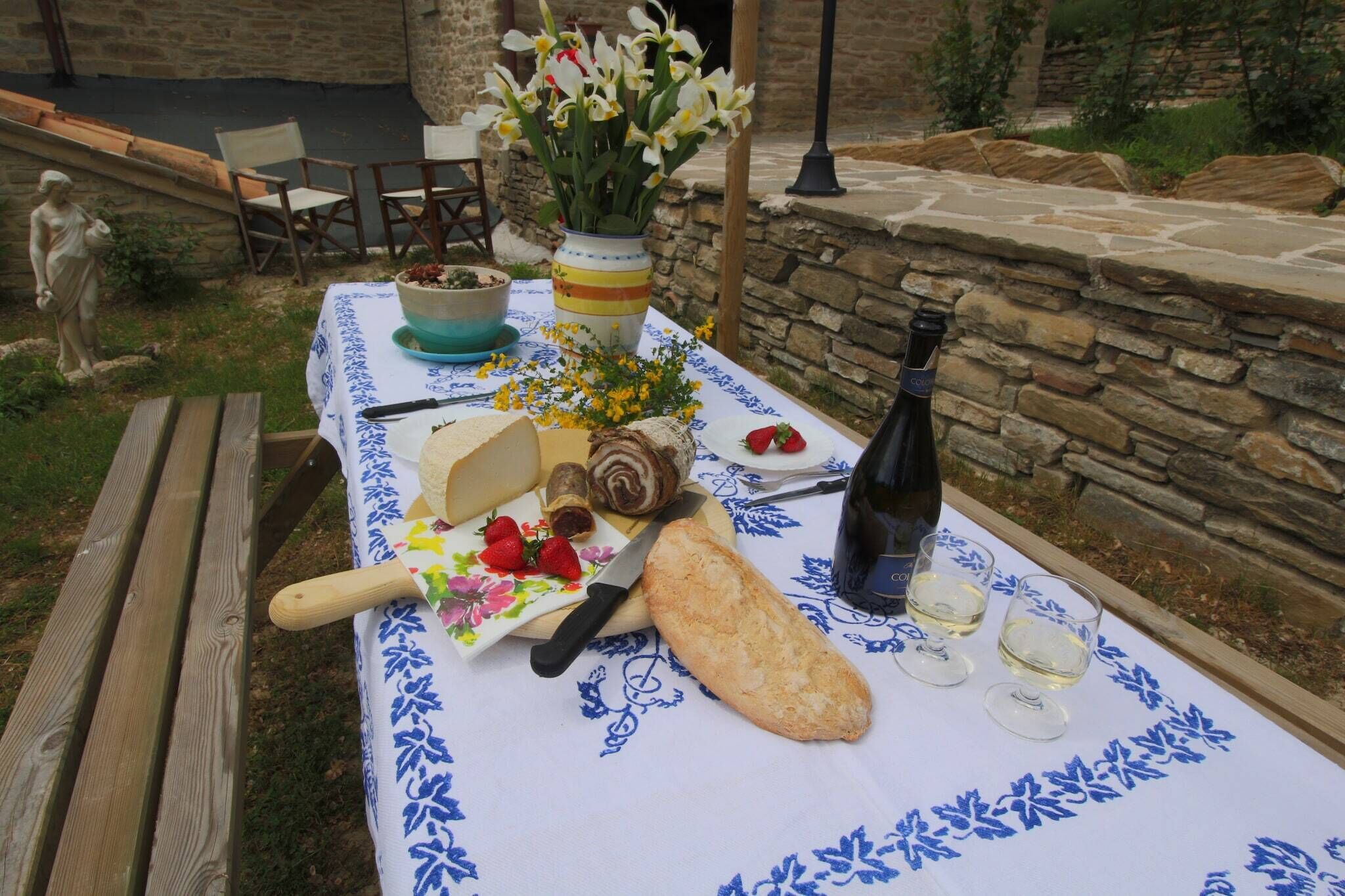 Un montaje para cenar al aire libre con una mesa dispuesta para dos personas, que incluye un pan, una botella de vino y un florero con flores.