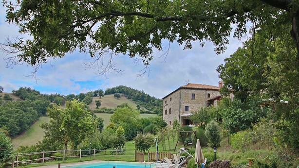 Zona de piscina con sillones de descanso, una mesa y un edificio de piedra al fondo.