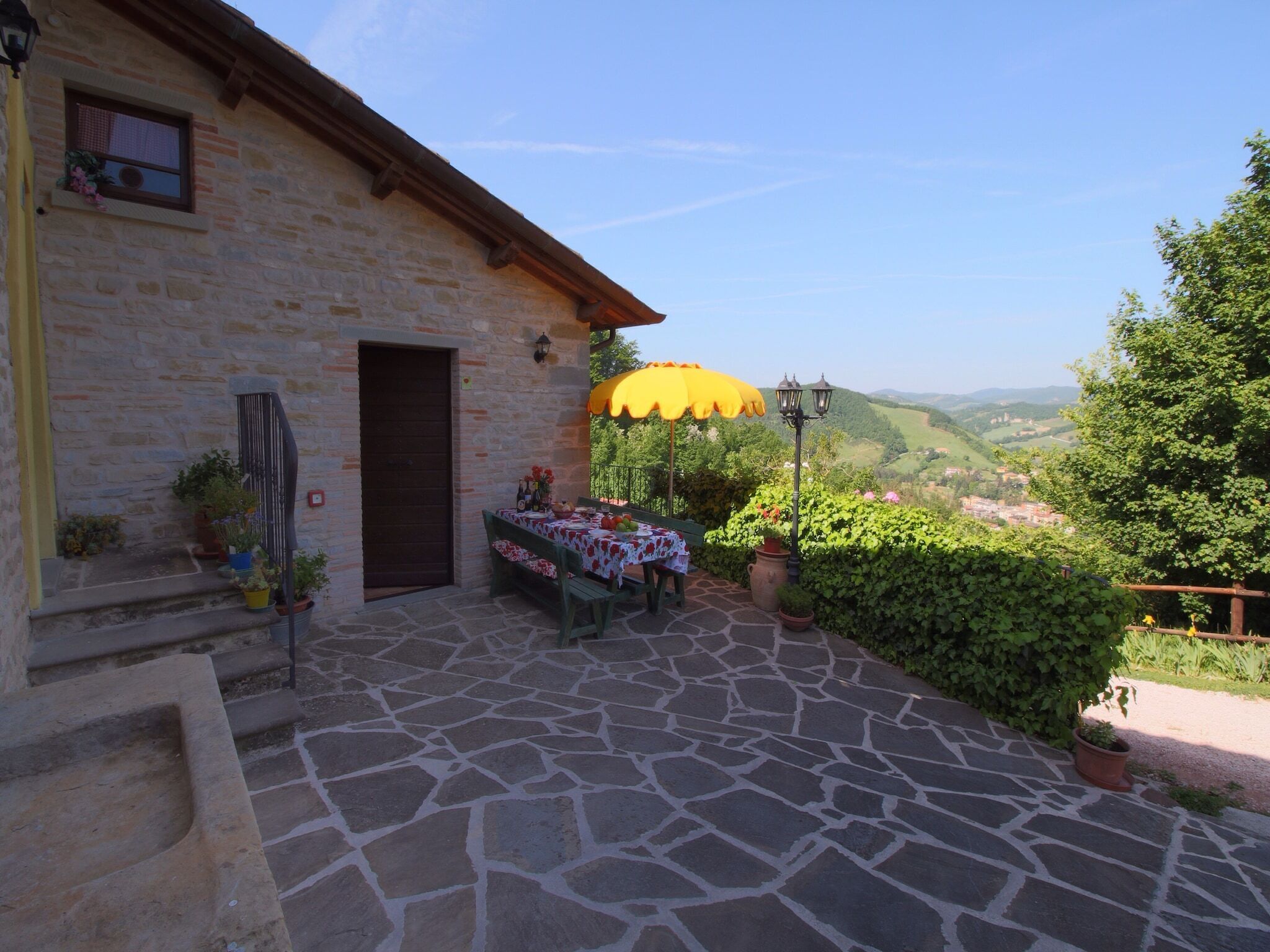 A stone building with a door and windows, a table with a tablecloth, potted plants, and a yellow umbrella.