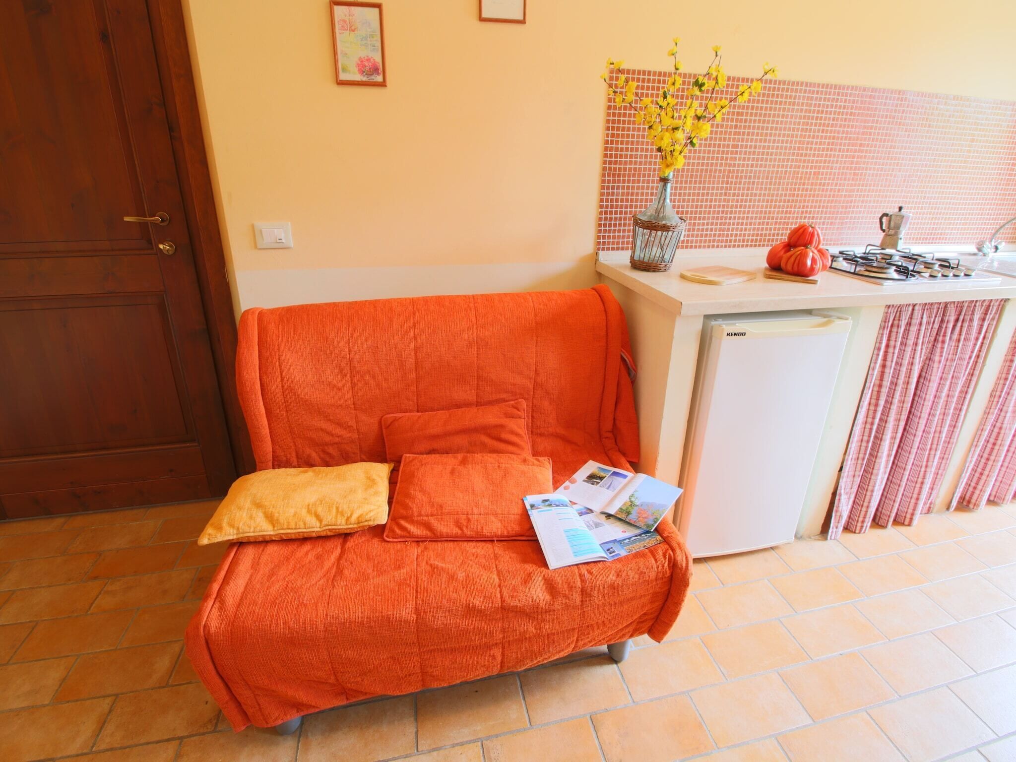 Een gezellige kamer met een enkele oranje bank, een klein wit keukenhoekje en een betegelde vloer.