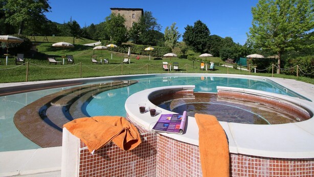 A swimming pool with a hot tub, surrounded by lounge chairs and umbrellas, with a castle in the background. - Apartment in Marche with Pool and Privacy (Apecchio)