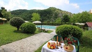 A poolside table with fruits, wine glasses, and a candle, surrounded by greenery and a pool. - Apartment in Marche with Pool and Privacy (Apecchio)