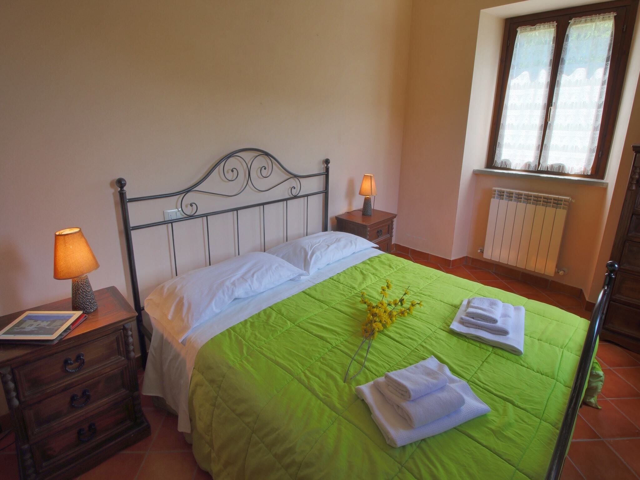 A neatly made bed with a green bedspread, two pillows, and a decorative branch. There are two bedside tables with lamps and a small drawer unit. A radiator is visible in the corner, and a window with curtains is on the right.