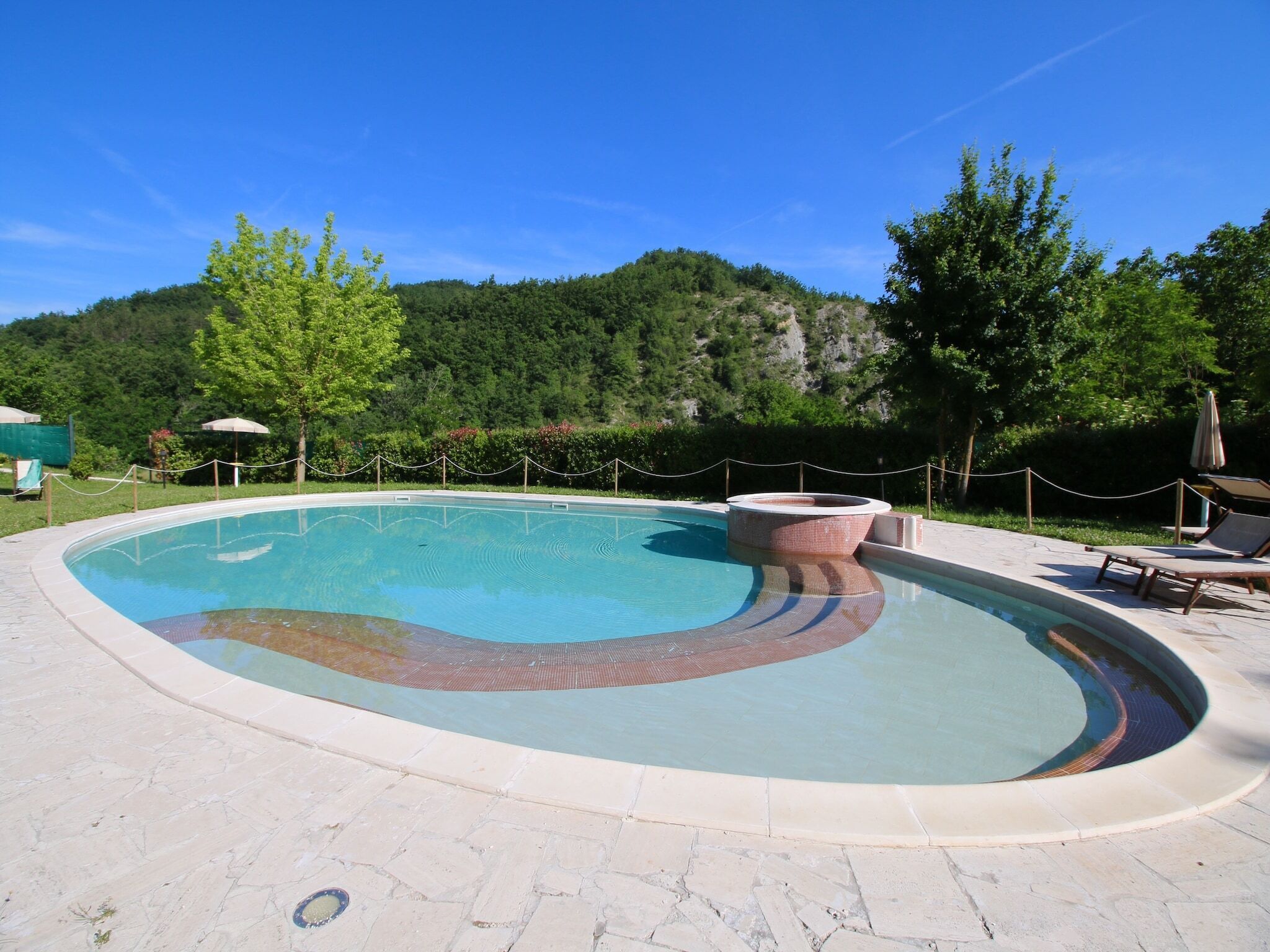 A swimming pool with a hot tub, surrounded by a stone deck and a rope barrier.