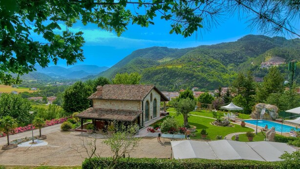 Um edifício de pedra com telhado em telhas, rodeado por um jardim bem cuidado com piscina, cascata e vista para as colinas ao redor.