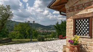Pátio com calçamento de pedra, um edifício rústico, uma janela decorativa e vista para uma cidade e montanhas ao longe.