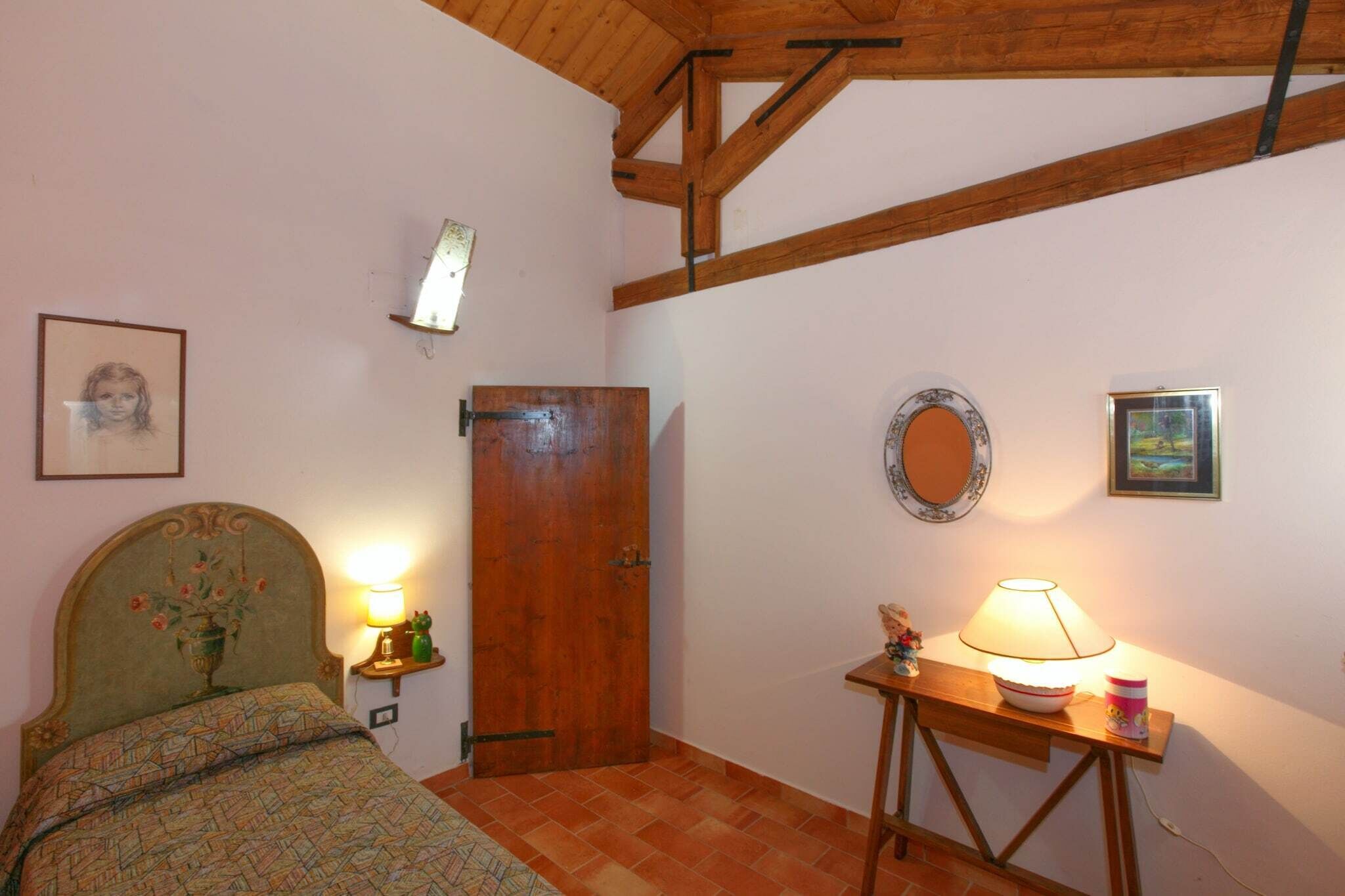 Ein Schlafzimmer mit einem Bett, einer Holztür, einem Nachttisch mit Lampe, einem Spiegel und einem gerahmten Bild an der Wand.